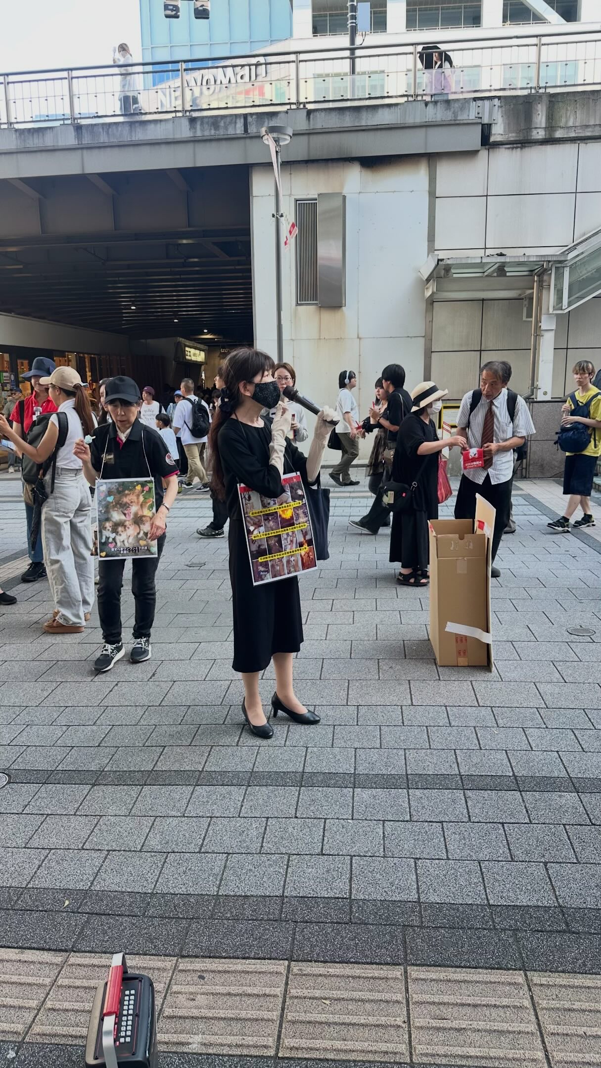 Eine Straßenaktion auf dem Vorplatz eines Bahnhofs, dessen Beschilderung auf Japanisch ist. Eine Person mit Maske steht in der Mitte, vor der Brust ein Schild mit Fotos von Wangwang und vom Geschehen; links trägt eine weitere Person ein Schild mit Fotos der Welpen. Passanten queren das Bild, dahinter eine Überführung und das Bahnhofsgebäude.