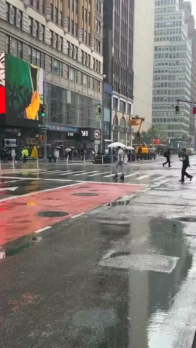 Eine New Yorker Straße im Regen: Passanten überqueren die nasse Fahrbahn, links eine rot markierte Fußgängerzone, dahinter Hochhäuser und Geschäftsschilder.
