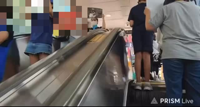 Neben einer Rolltreppe in einem Innenraum hält jemand eine rote Unterstützungsbotschaft; alle sichtbaren Gesichter sind in der öffentlichen Fassung verdeckt.