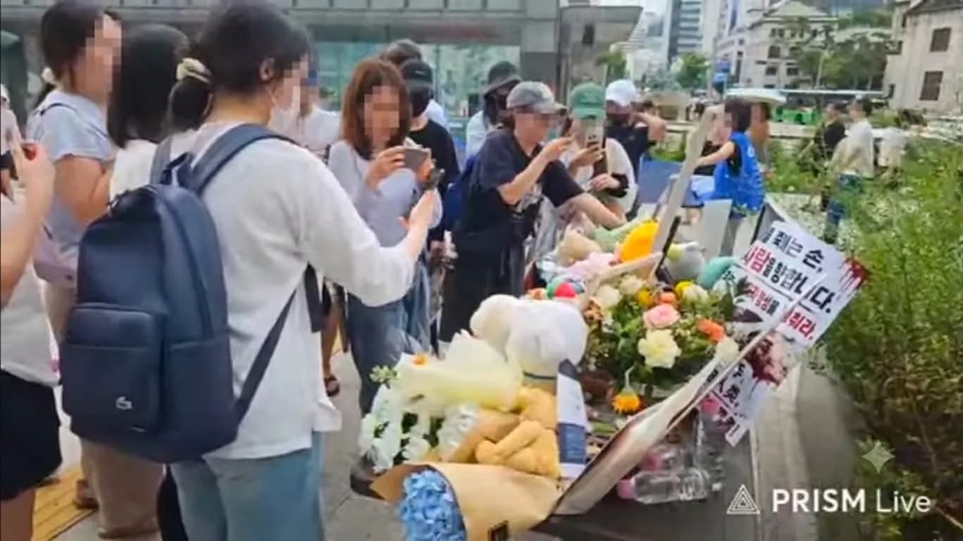 Eine Gedenkversammlung auf der Straße, die Schilder sind auf Koreanisch. Teilnehmende stehen am Gehweg, einige halten ihre Handys hoch, um die Blumen, Stofftiere und Schilder am Boden zu fotografieren. Alle erkennbaren Gesichter wurden im Originalbild verpixelt.